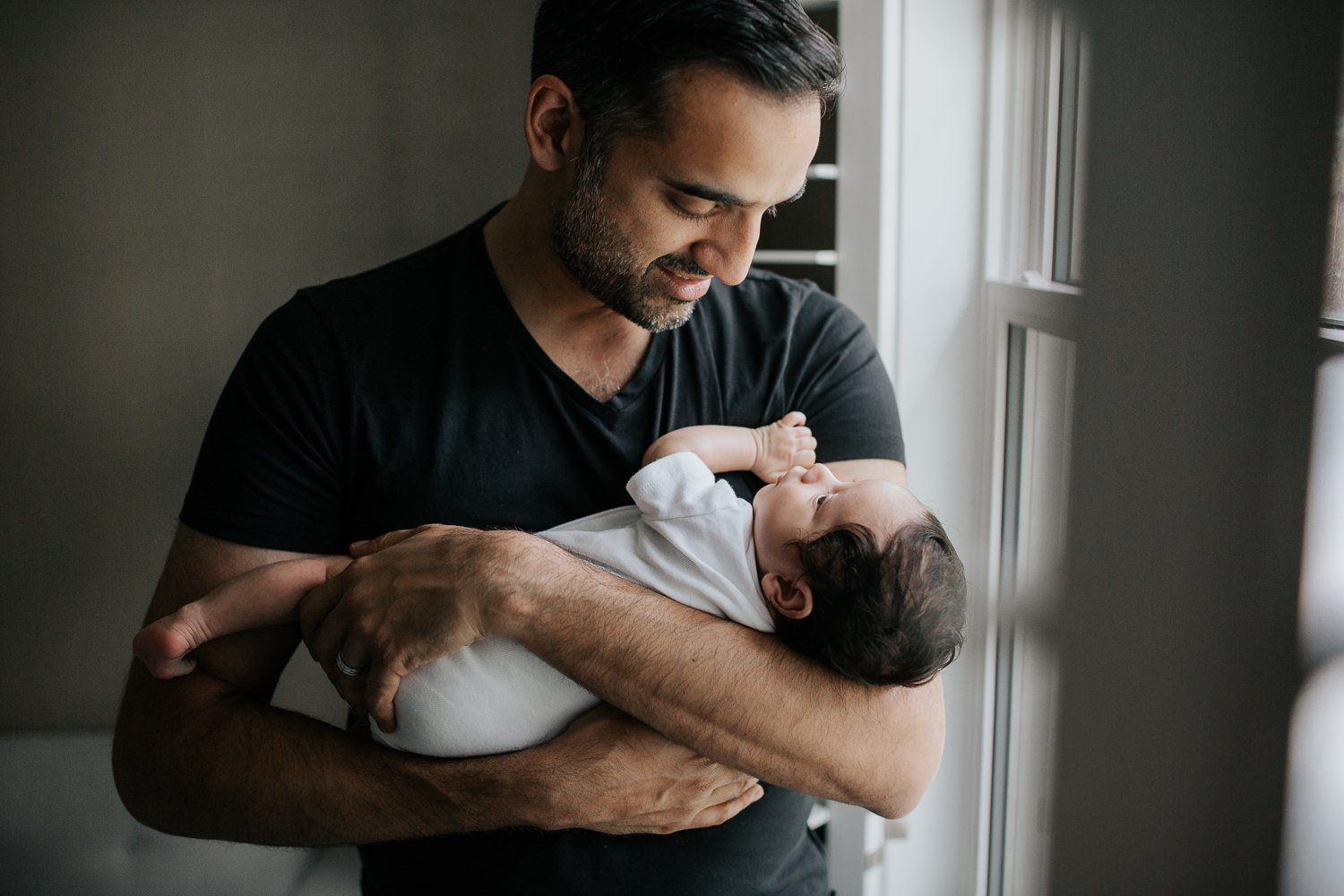 new father standing in front of window holding and smiling at 1 month old baby boy in onesie in his arms - Markham In-Home Photos
