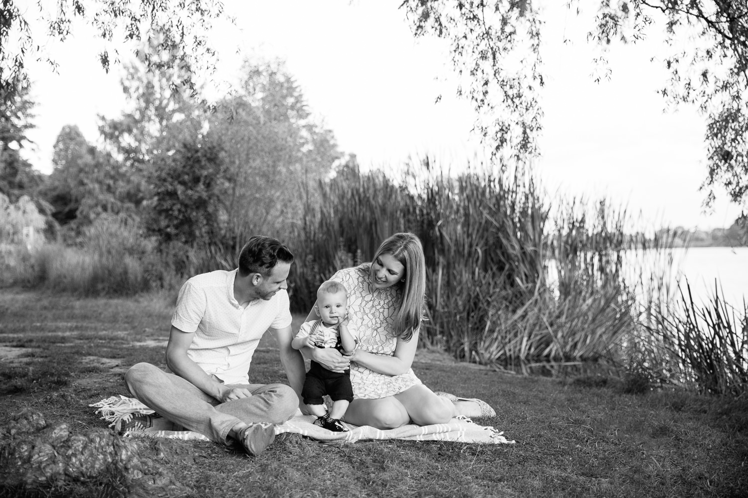 family of 3 sitting on blanket under large willow tree next to pond at high park, 8 month old baby boy standing between mom and dad looking at camera - Stouffville Lifestyle Photography