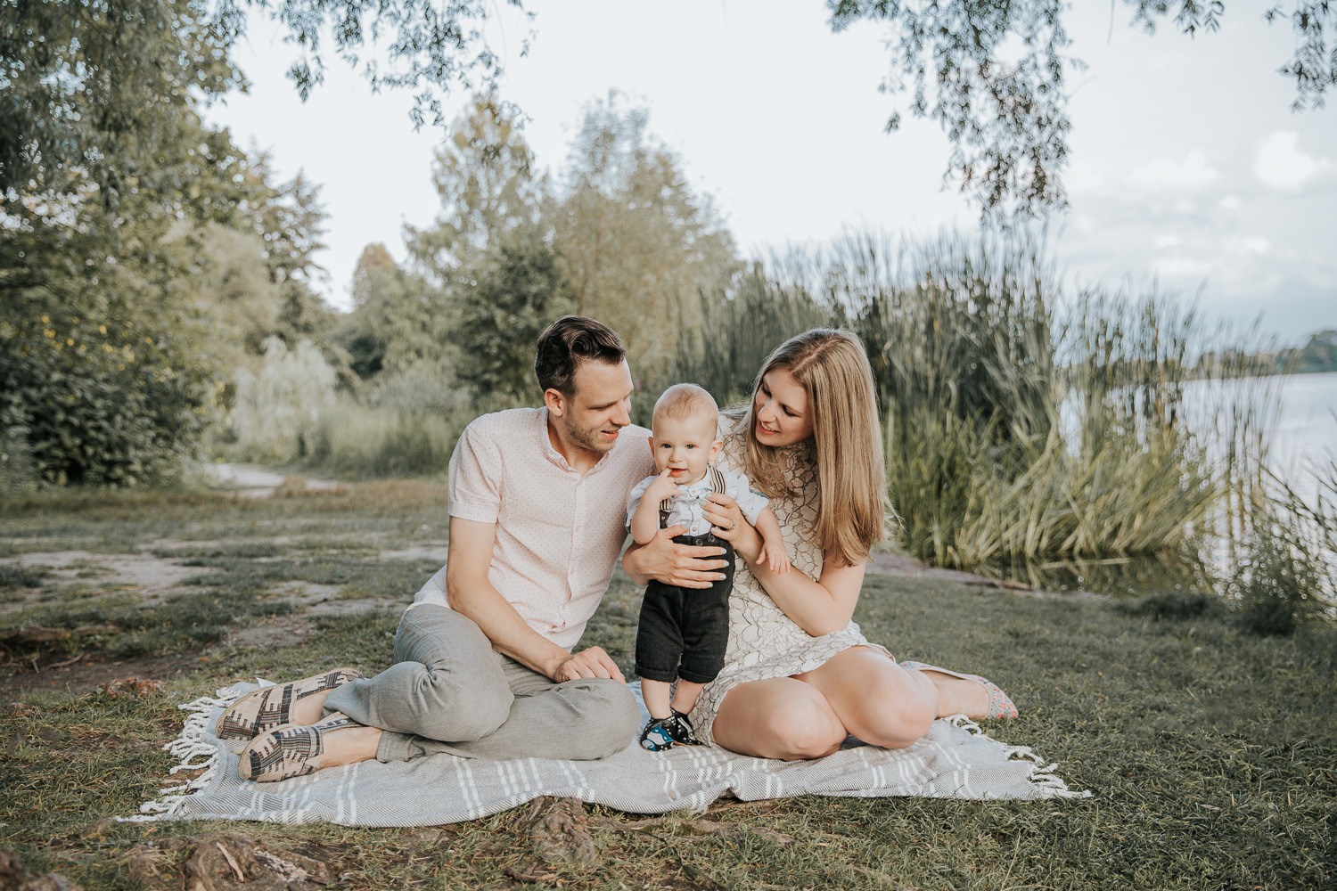 family of 3 sitting on blanket under large willow tree next to pond at high park, 8 month old baby boy standing between mom and dad looking at camera - Markham Lifestyle Photography