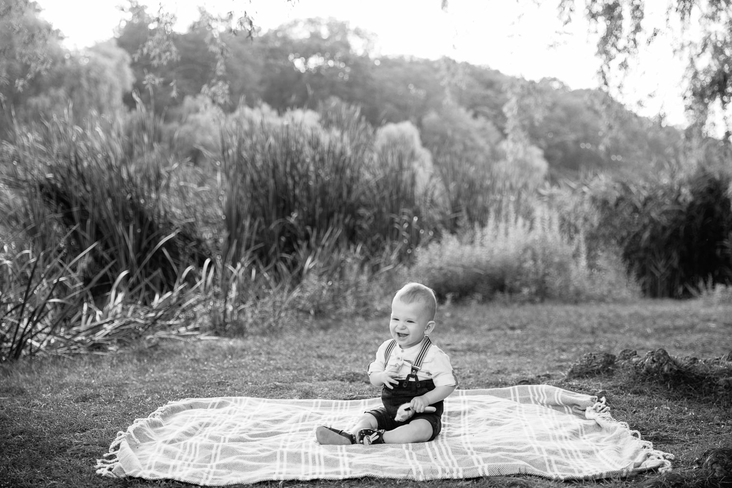 8 month old baby boy sitting on blanket on grass at high park wearing suspenders and button down shirt, holding toy and laughing - Markham In-Home Photography