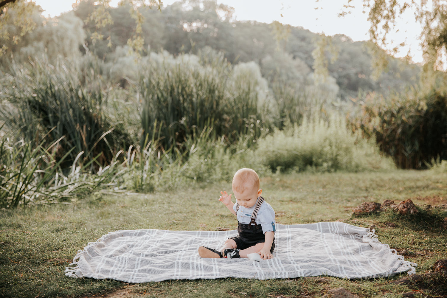 8 month old baby boy sitting on blanket on grass at high park wearing suspenders and button down shirt, looking down playing with toy - Stouffville In-Home Photography