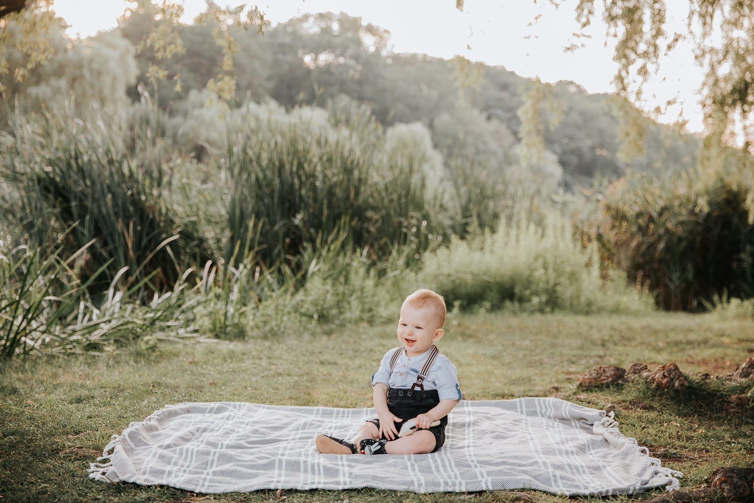 8 month old baby boy sitting on blanket on grass at high park wearing suspenders and button down shirt, holding toy and smiling - Barrie In-Home Photography