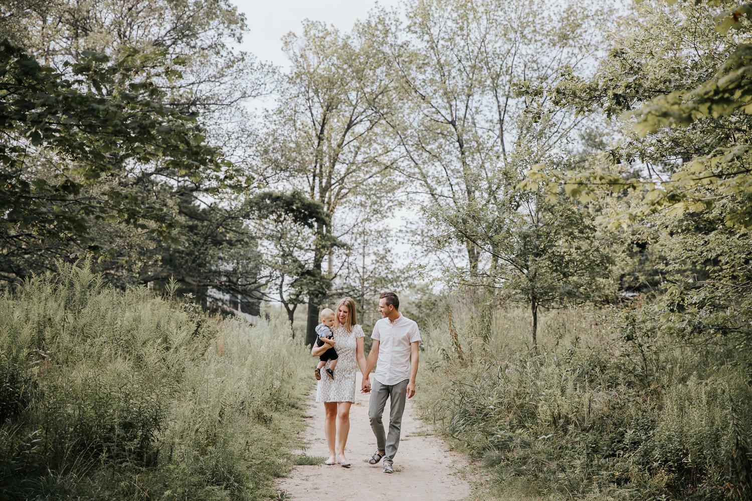 family of 3 walking down dirt path at high park surrounded by tall grasses and trees, mom carrying 8 month old blonde baby boy and parents holding hands - GTA In-Home Photography