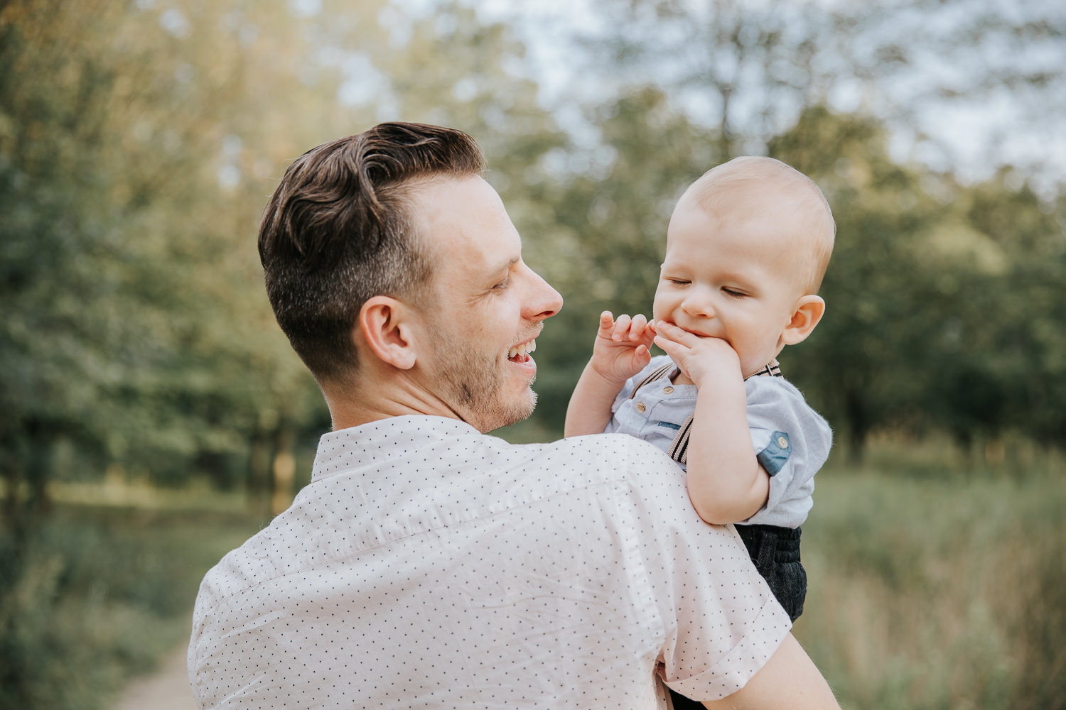 father holding 8 month old blonde baby boy, son is chewing hand and father is laughing at him, in high park - Markham In-Home Photos