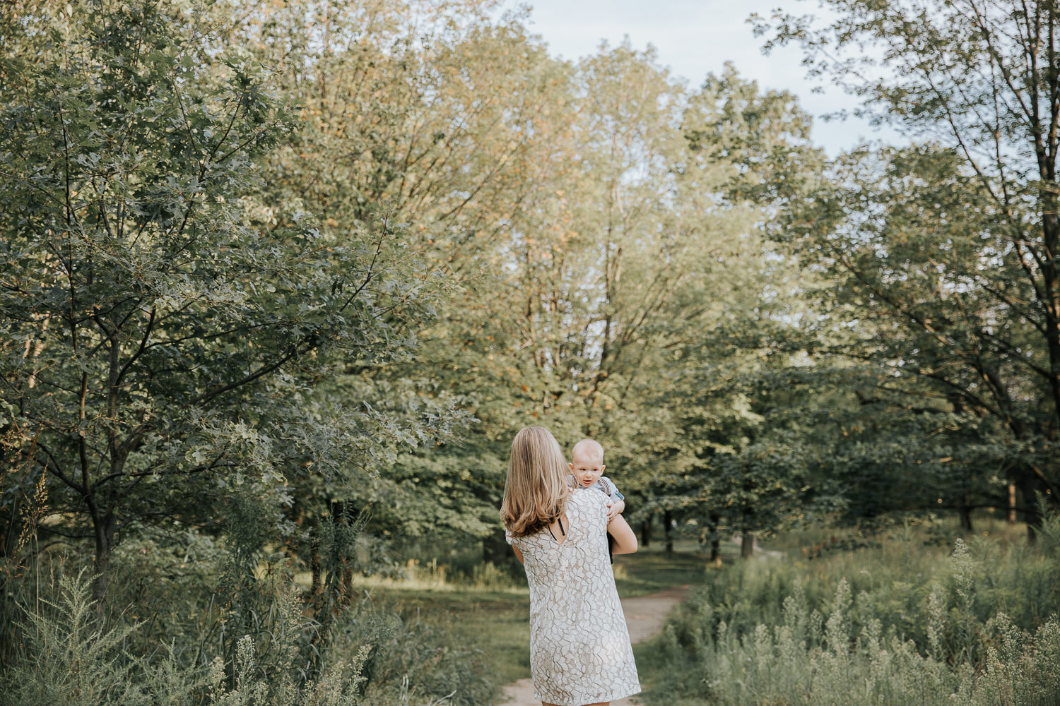 mother carrying 7 month old blonde baby boy down path at high park surrounded by greenery, son peeking over mom's shoulder - Stouffville In-Home Photos