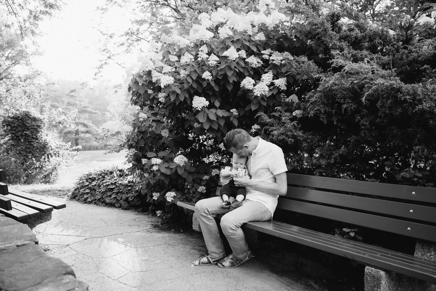 dad sitting on bench in garden at high park with 8 month old blonde baby boy in his lap, snuggling and playing with toy - Markham Lifestyle Photos