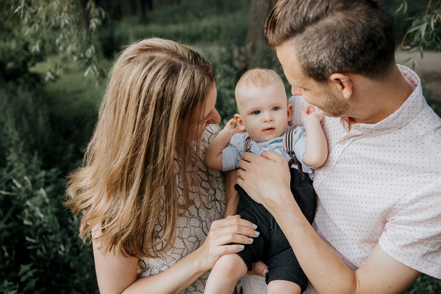 family of 3 standing under willow tree next to pond at high park, father carrying 8 month old blonde baby boy and mom with hand on his leg, parents smiling at son - Newmarket Lifestyle Photography