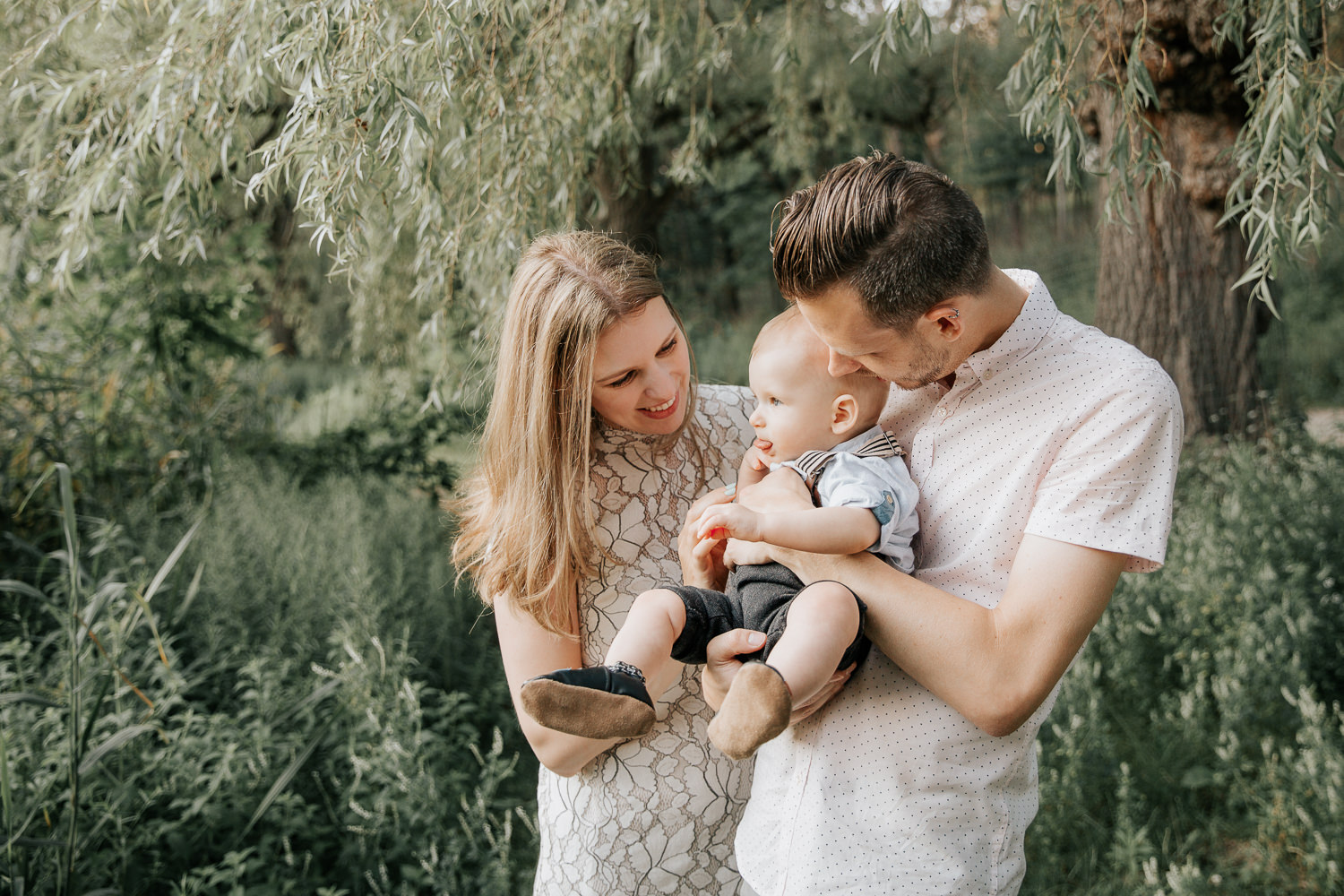 family of 3 standing under willow tree next to pond at high park, father carrying 8 month old blonde baby boy and mom holding his hand, parents smiling at son - York Region Lifestyle Photos