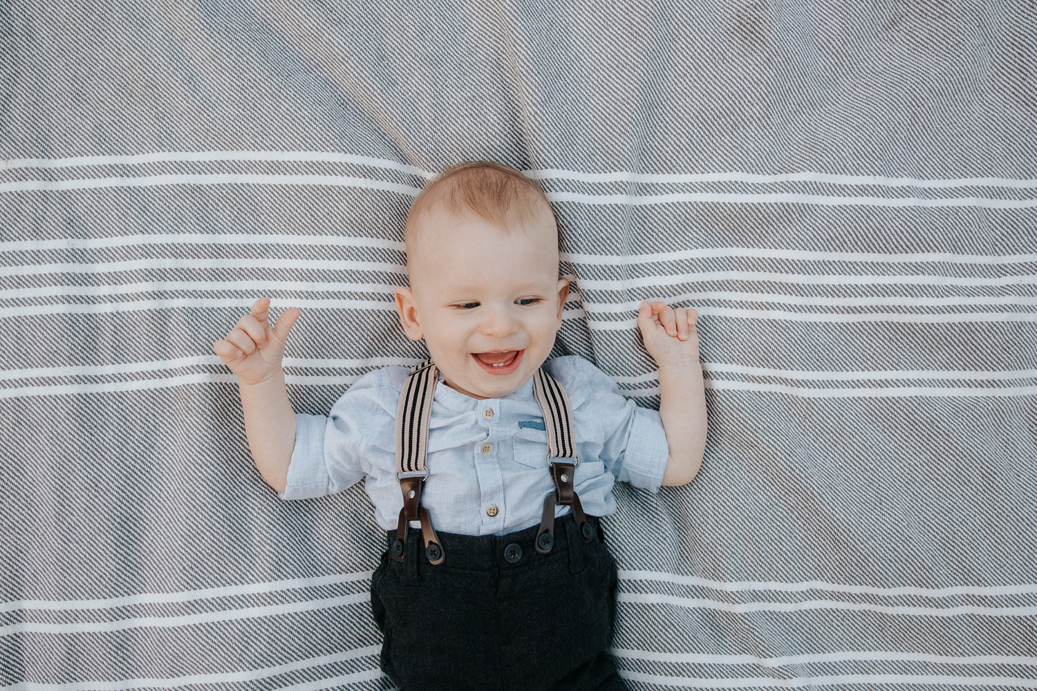 8 month old baby boy lying on grey and white striped blanket, arms in the air and laughing showing two bottom teeth - GTA Lifestyle Photography