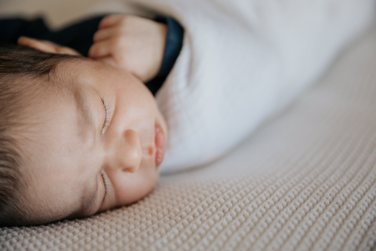 2 week old baby boy with dark hair in navy blue onesie wrapped in white swaddle, sleeping on bed, shot of eyelashes - York Region Lifestyle Photography