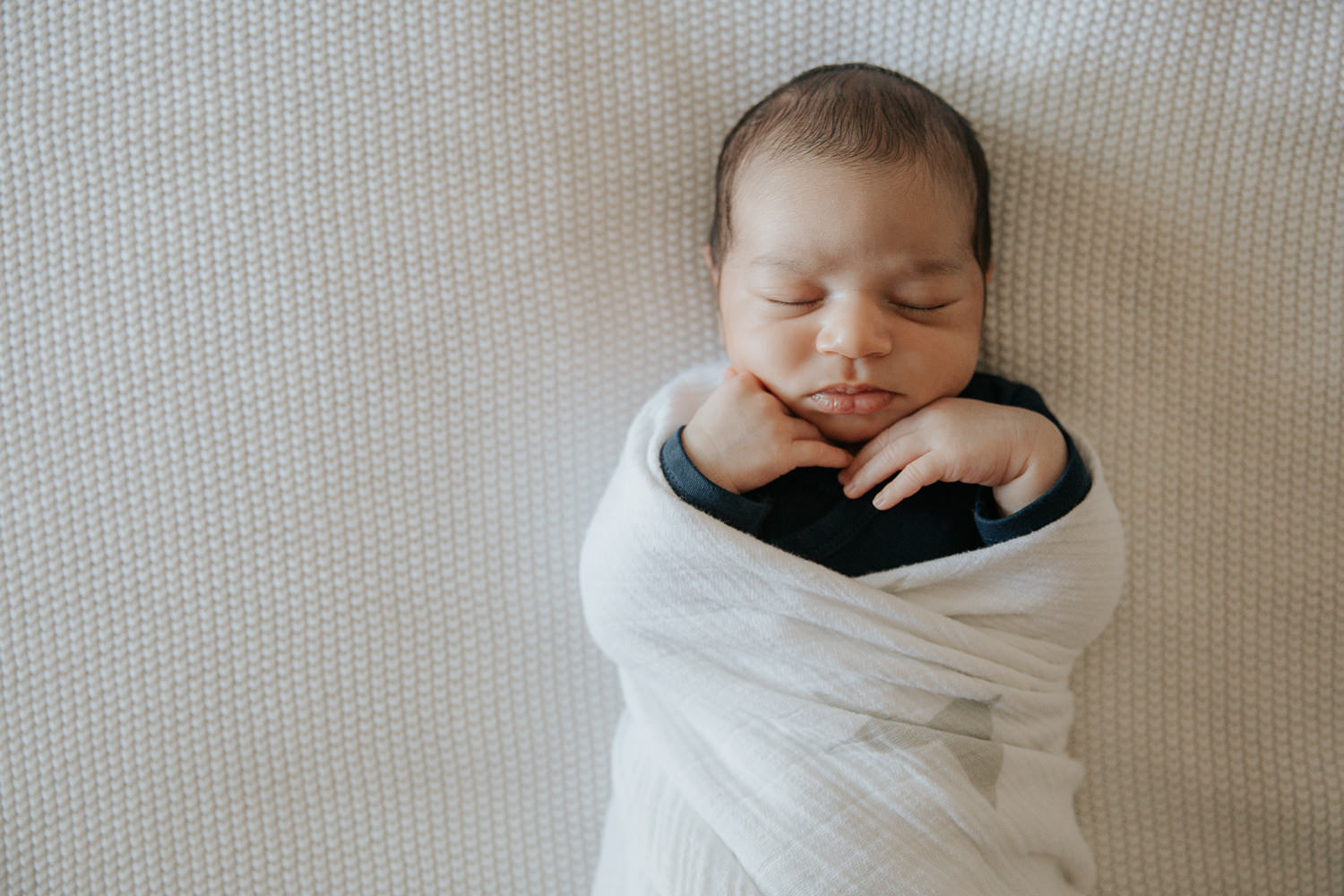 2 week old baby boy with dark hair in navy blue onesie wrapped in white swaddle, sleeping on bed with hands tucked under chin - Markham Lifestyle Photography