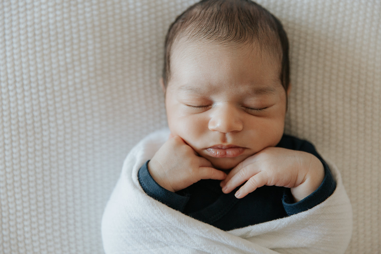 2 week old baby boy with dark hair in navy blue onesie wrapped in white swaddle, sleeping on bed with hands tucked under chin - Newmarket Lifestyle Photography