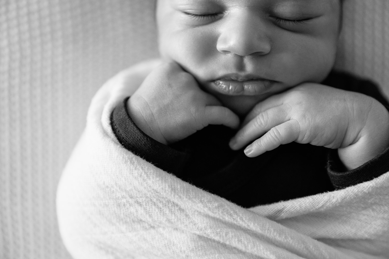 2 week old baby boy with dark hair in dark onesie wrapped in white swaddle, sleeping on bed with hands tucked under chin - Stouffville Lifestyle Photography