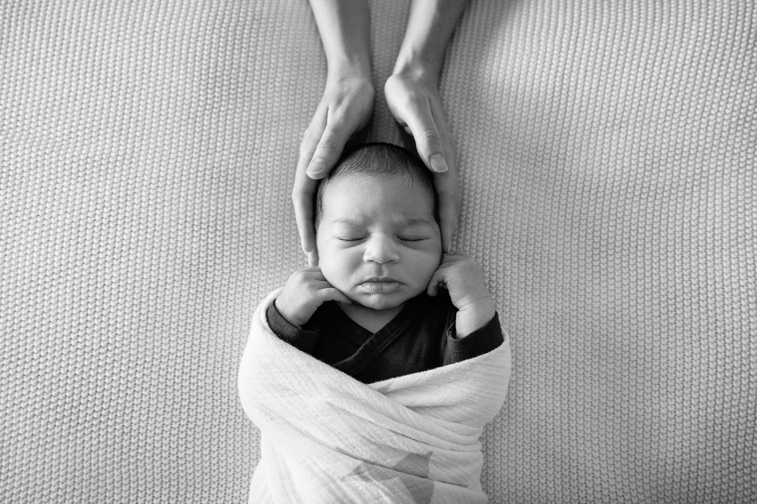 2 week old baby boy with dark hair in dark onesie wrapped in white swaddle, sleeping on bed with hands beside cheeks, mom's hands cradling son's head - Stouffville In-Home Photography