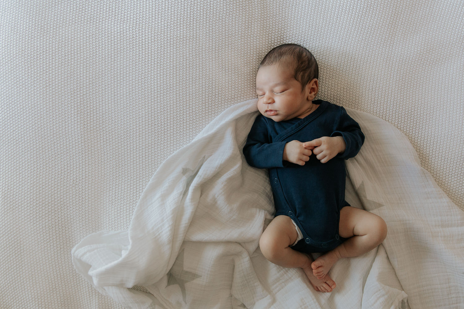 2 week old baby boy with dark hair and olive skin wearing navy blue onesie sleeping on swaddle on bed, hands and legs tucked in - Markham In-Home Photography