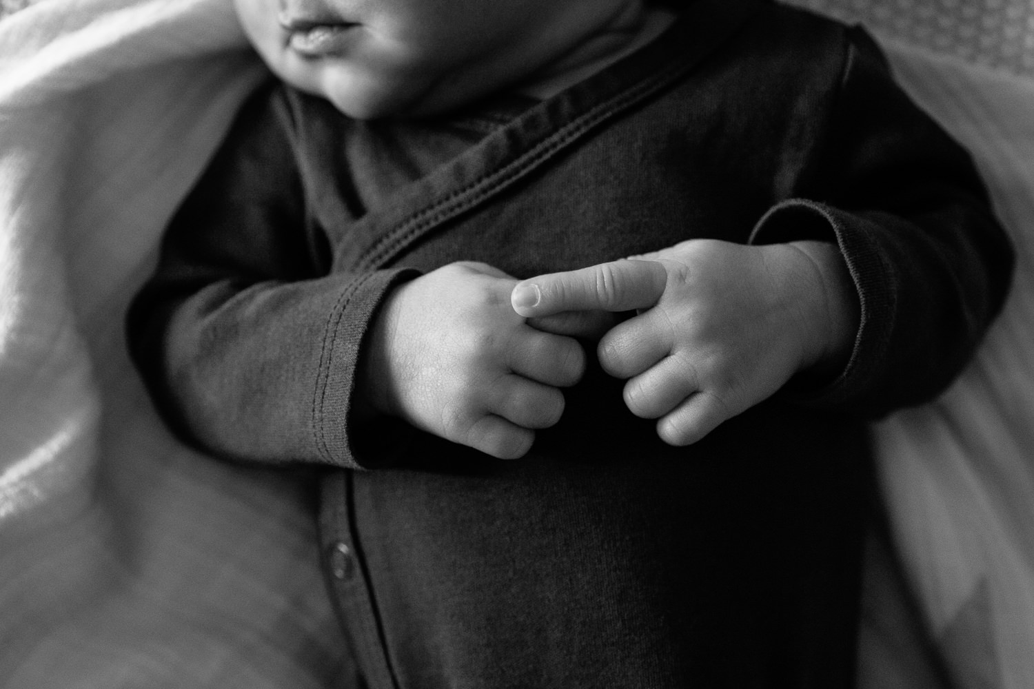 2 week old baby boy wearing dark onesie lying on bed, close up of hands clasped on chest - Barrie In-Home Photography