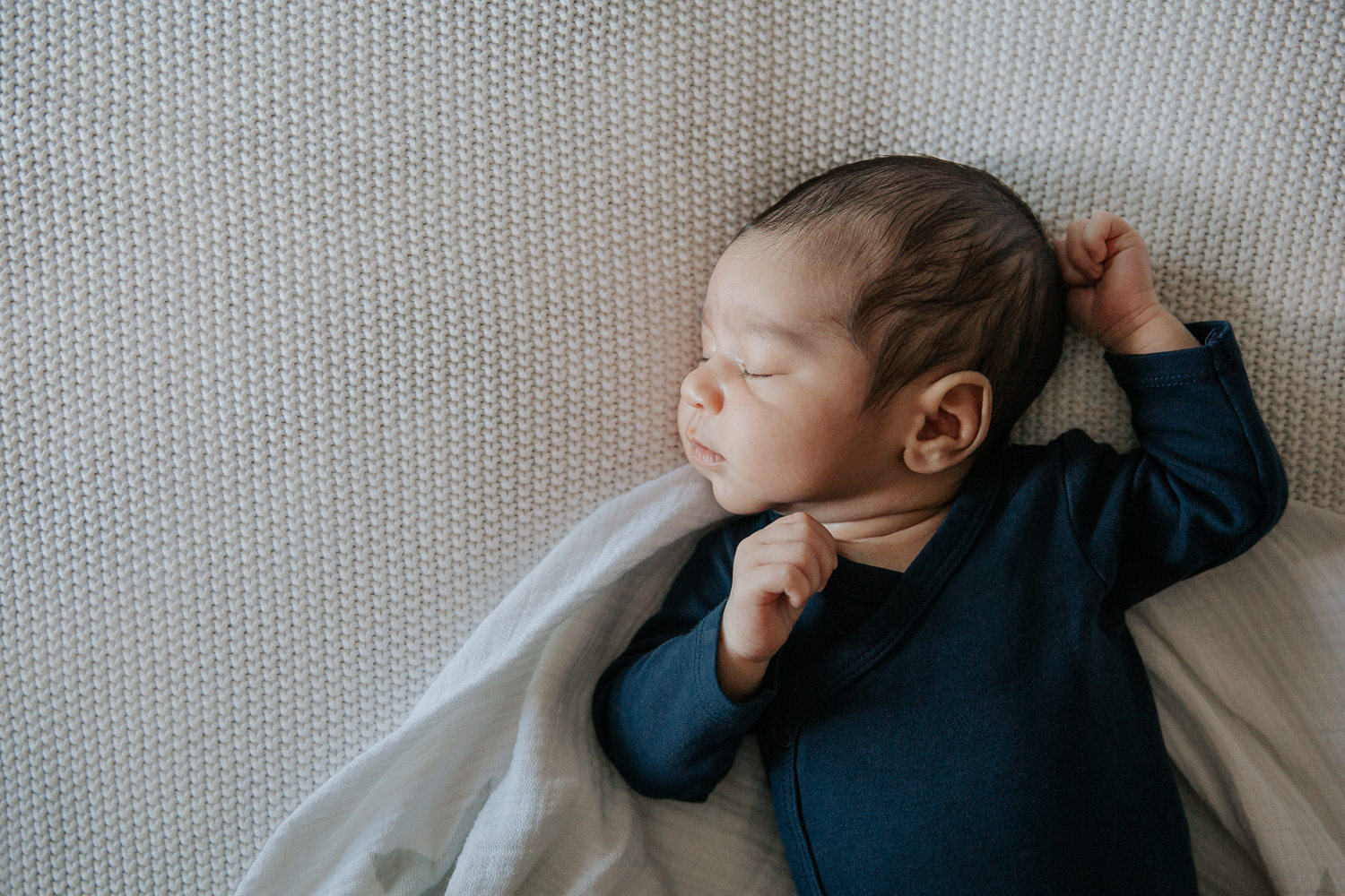 2 week old baby boy with dark hair and olive skin wearing navy blue onesie sleeping on swaddle on bed, hands up around head - York Region In-Home Photography