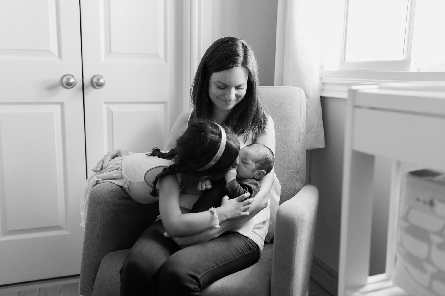 mom sitting in grey glider in neutral nursery holding 2 week old baby boy as 4 year old big sister leans over and kisses brother - York Region In-Home Photos
