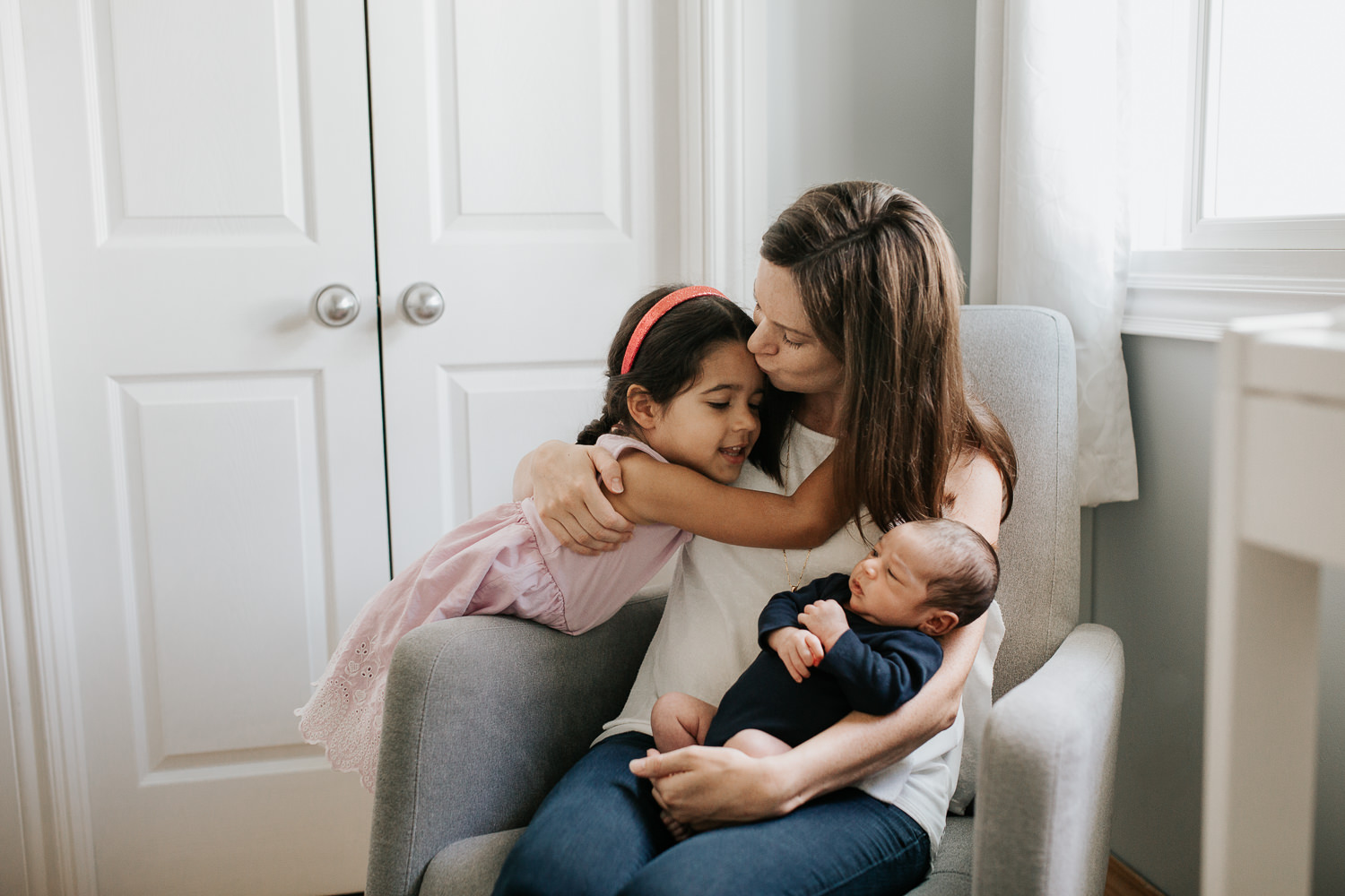 mom with long brown hair in white top and jeans sitting in grey glider in neutral nursery holding 2 week old baby boy and kissing 4 year old daughter on forehead - GTA In-Home Photos