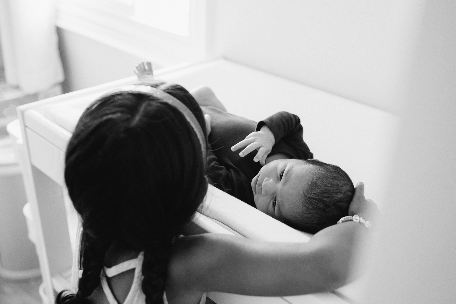 4 year old toddler girl with dark hair in braids peeking over to see 2 week old baby brother lying on nursery change table - Barrie Lifestyle Photos