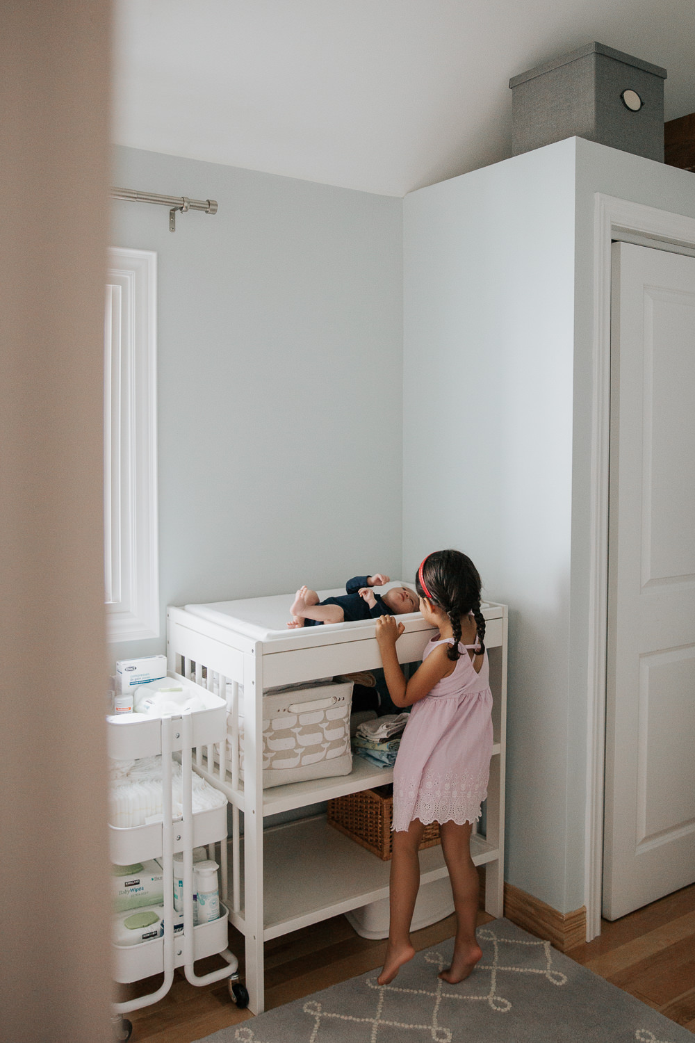 4 year old toddler girl with dark hair and braids in purple dress standing on tiptoes to see 2 week old baby brother lying on nursery change table - Markham Lifestyle Photos