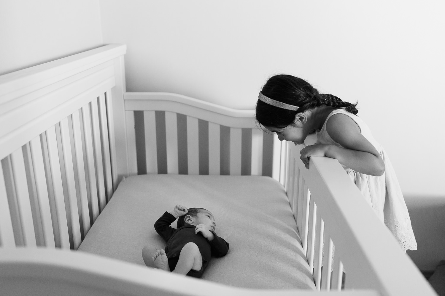 4 year old toddler girl with dark hair in braids peeking over to see 2 week old baby brother lying in crib in nursery - GTA Lifestyle Photos