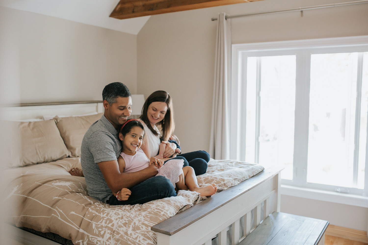 family of four sitting on bed, 4 year old toddler girl sitting in dad's lap smiling at camera, mom holding 2 week old baby boy and smiling at daughter - Markham Lifestyle Photography
