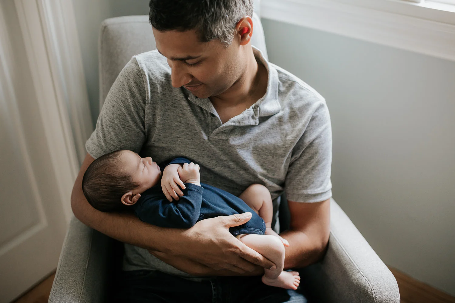father sitting in nursery rocker holding 2 week old baby boy in navy blue onesie who is sleeping with arms crossed - GTA Lifestyle Photography