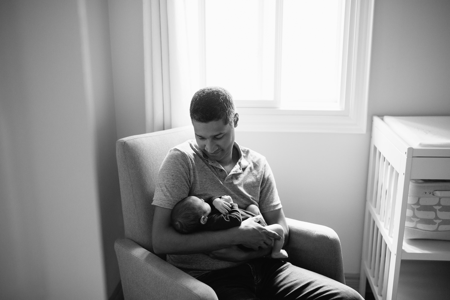 father sitting in nursery rocker holding 2 week old baby boy in navy blue onesie who is sleeping with arms crossed - York Region Lifestyle Photography