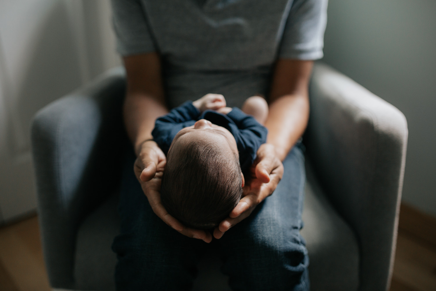 father sitting in nursery rocker holding 2 week old baby son in navy blue onesie who is sleeping with arms crossed, close up of son's head in dad's hands - Newmarket In-Home Photography