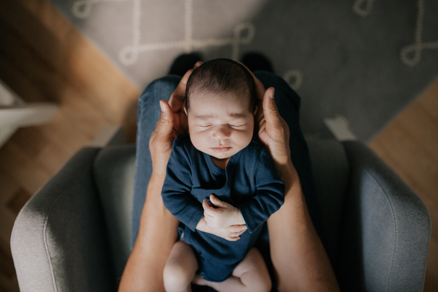 father sitting in nursery rocker holding 2 week old baby boy in navy blue onesie who is sleeping with arms crossed, shot from above - Stouffville In-Home Photography