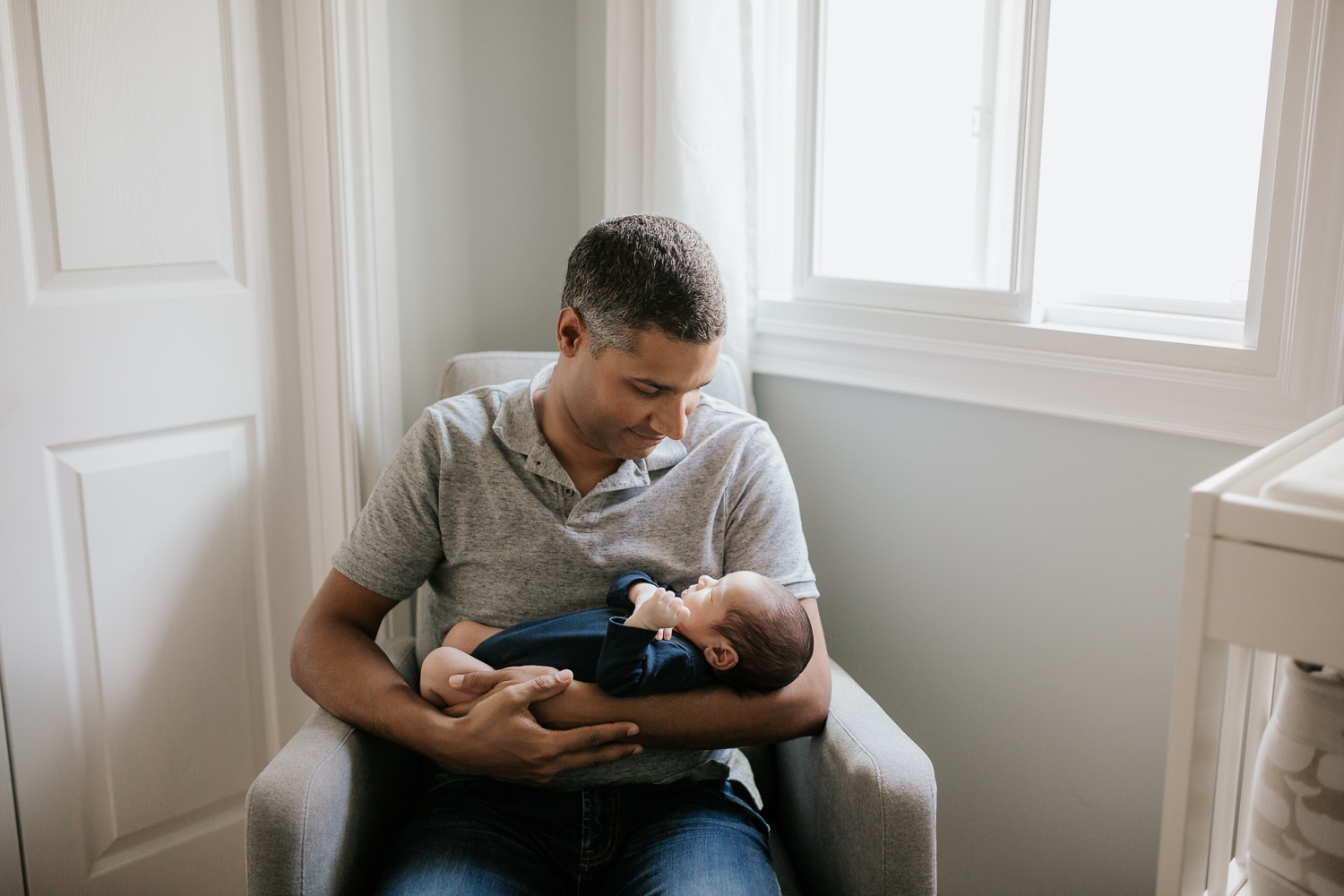 father sitting in nursery rocker holding 2 week old baby boy in navy blue onesie who is sleeping - Markham In-Home Photography