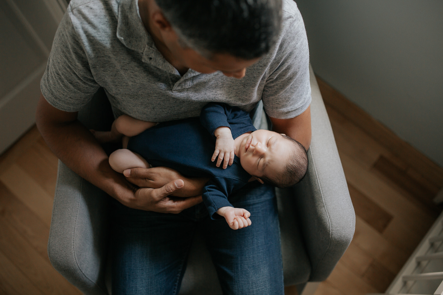 father sitting in nursery rocker holding 2 week old baby boy in navy blue onesie who is sleeping, shot from above - GTA In-Home Photography