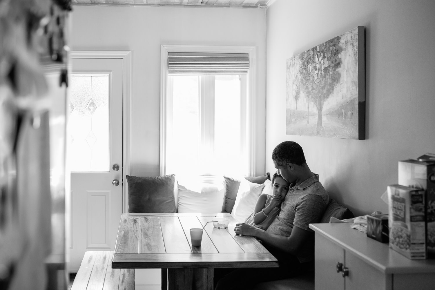 4 year old toddler girl with dark hair sitting at kitchen table with dad eating a snack and looking seriously at the camera - York Region In-Home Photos