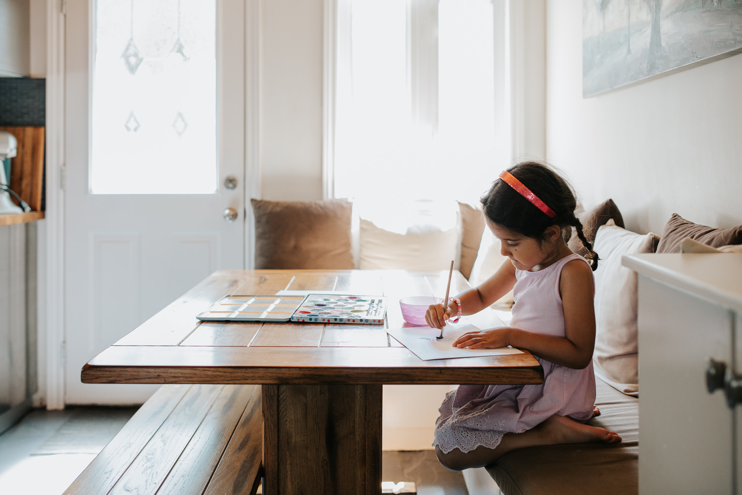 4 year old toddler girl with dark hair in pigtails wearing purple dress sitting at kitchen table painting with watercolours - Newmarket Lifestyle Photos