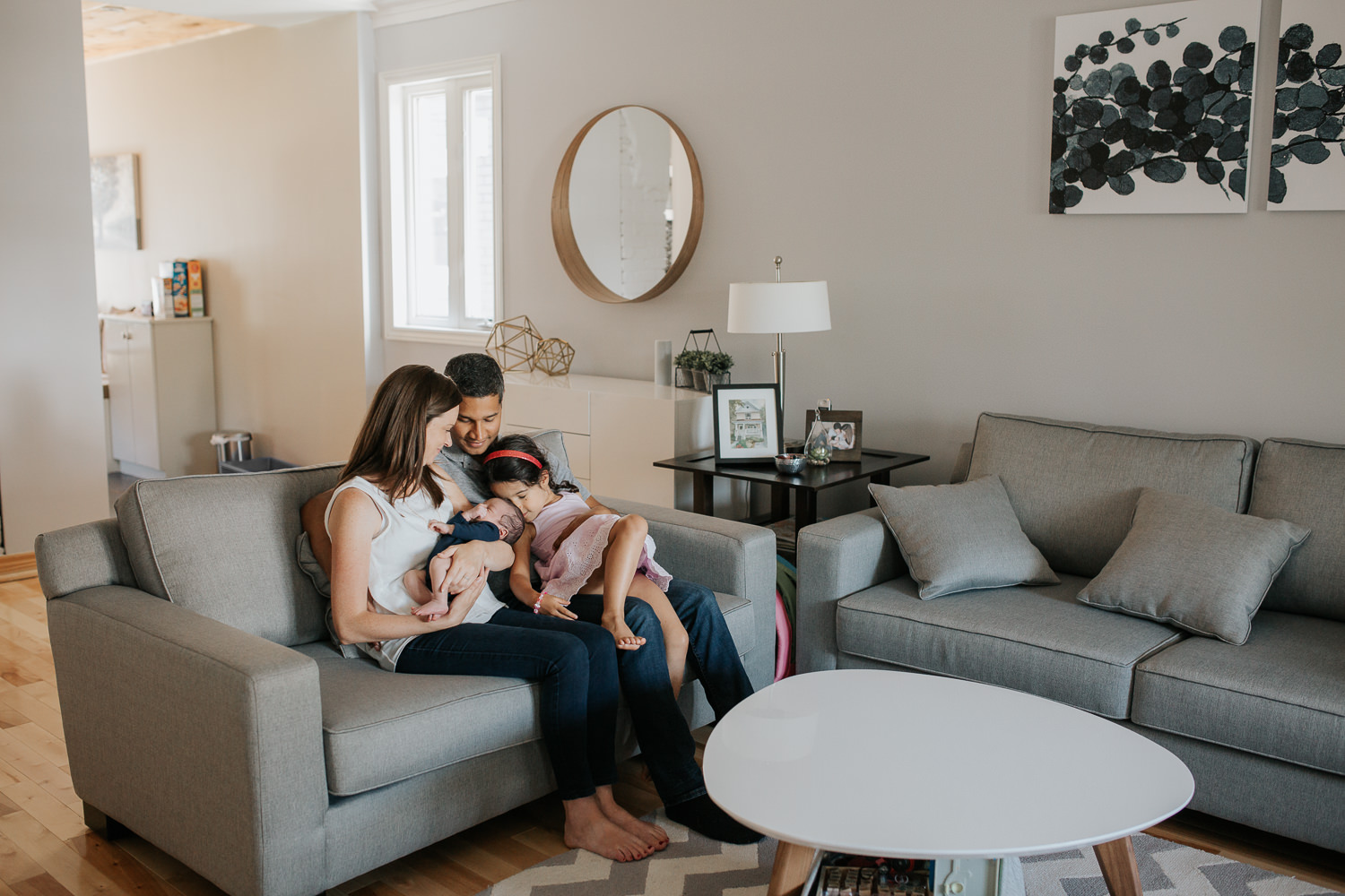 family of 4 sitting on living room couch, 4 year old toddler girl sitting in dad's lap and leaning face against 2 week old baby brother's head who is being held in mom's arms - Barrie Lifestyle Photos