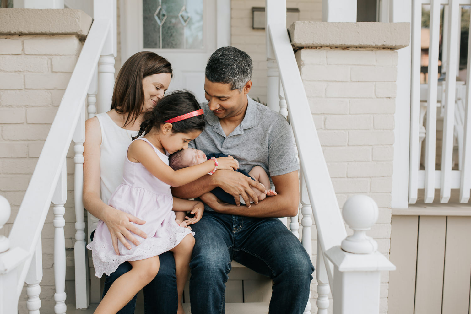 family of 4 sitting on front porch, dad holding 2 week old baby boy in his arms and 4 year old toddler girl sitting in mom's lap and kissing little brother on head - Newmarket Lifestyle Photography