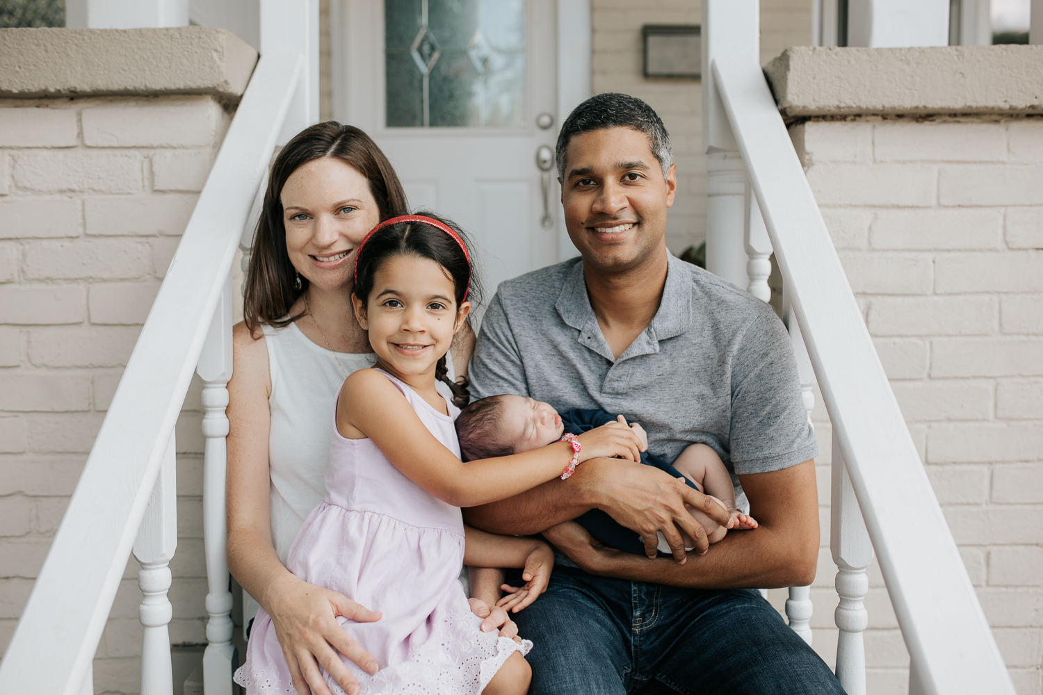family of 4 sitting on front porch looking at camera, dad holding 2 week old baby boy in his arms and 4 year old toddler girl sitting in mom's lap - York Region Lifestyle Photos