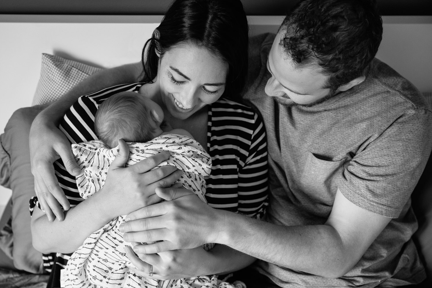family of 3, new parents cuddle on bed, mom holding 2 week old baby girl covered with black and white swaddle, dad embracing wife and daughter - Markham Lifestyle Photography