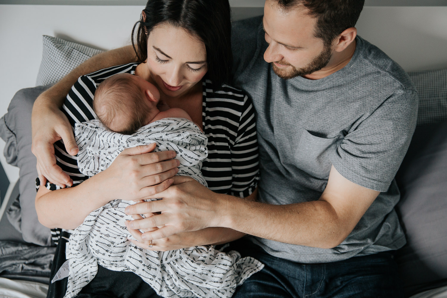 family of 3, new parents cuddle on bed, mom holding 2 week old baby girl covered with black and white swaddle, dad embracing wife and daughter - Stouffville Lifestyle Photography