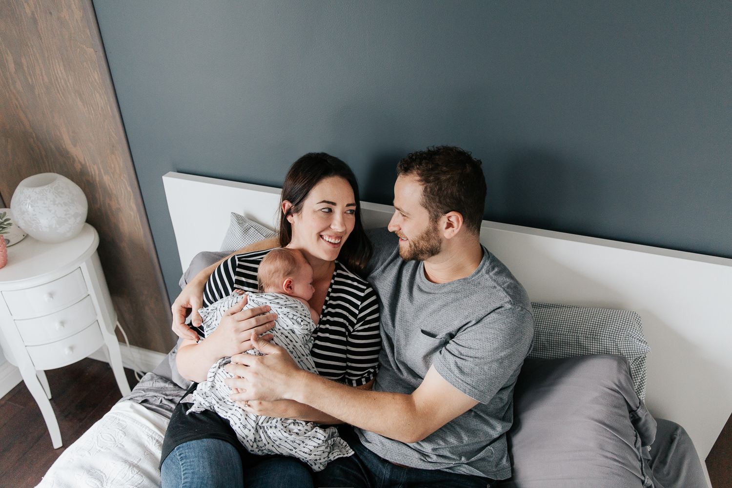 family of 3, new parents cuddle on bed smiling at each other, mom holding 2 week old baby girl covered with black and white swaddle, dad embracing wife and daughter - Barrie Lifestyle Photography