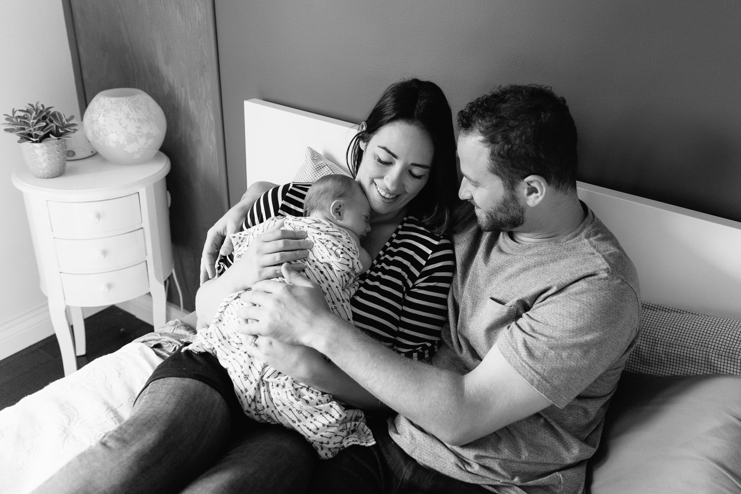 family of 3, new parents cuddle on bed, mom holding 2 week old baby girl covered with black and white swaddle, dad embracing wife and daughter - Uxbridge Lifestyle Photography