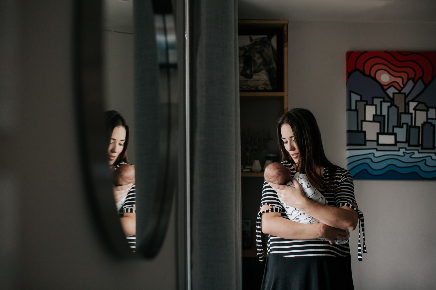 new mom in striped top with long dark brown hair standing next to window holding 2 week old baby girl with light hair who is sleeping in her arms, comforting daughter - Markham In-Home Photography