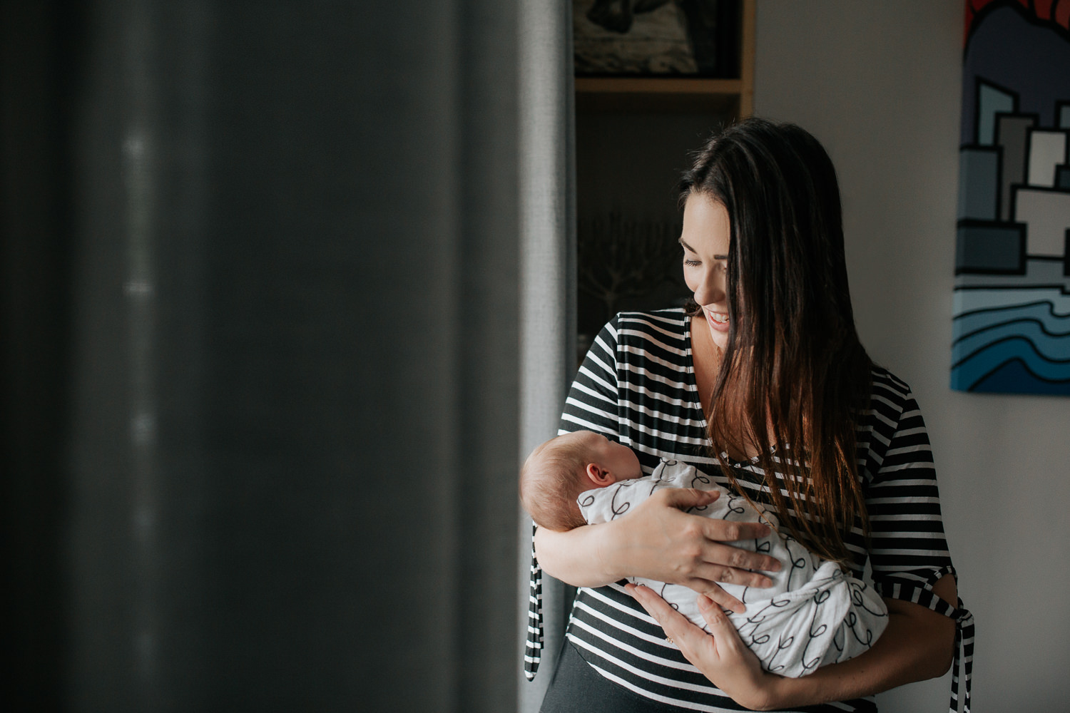 new mom in striped top with long dark brown hair standing next to window holding 2 week old baby girl with light hair who is sleeping in her arms, smiling at daughter - Stouffville In-Home Photography