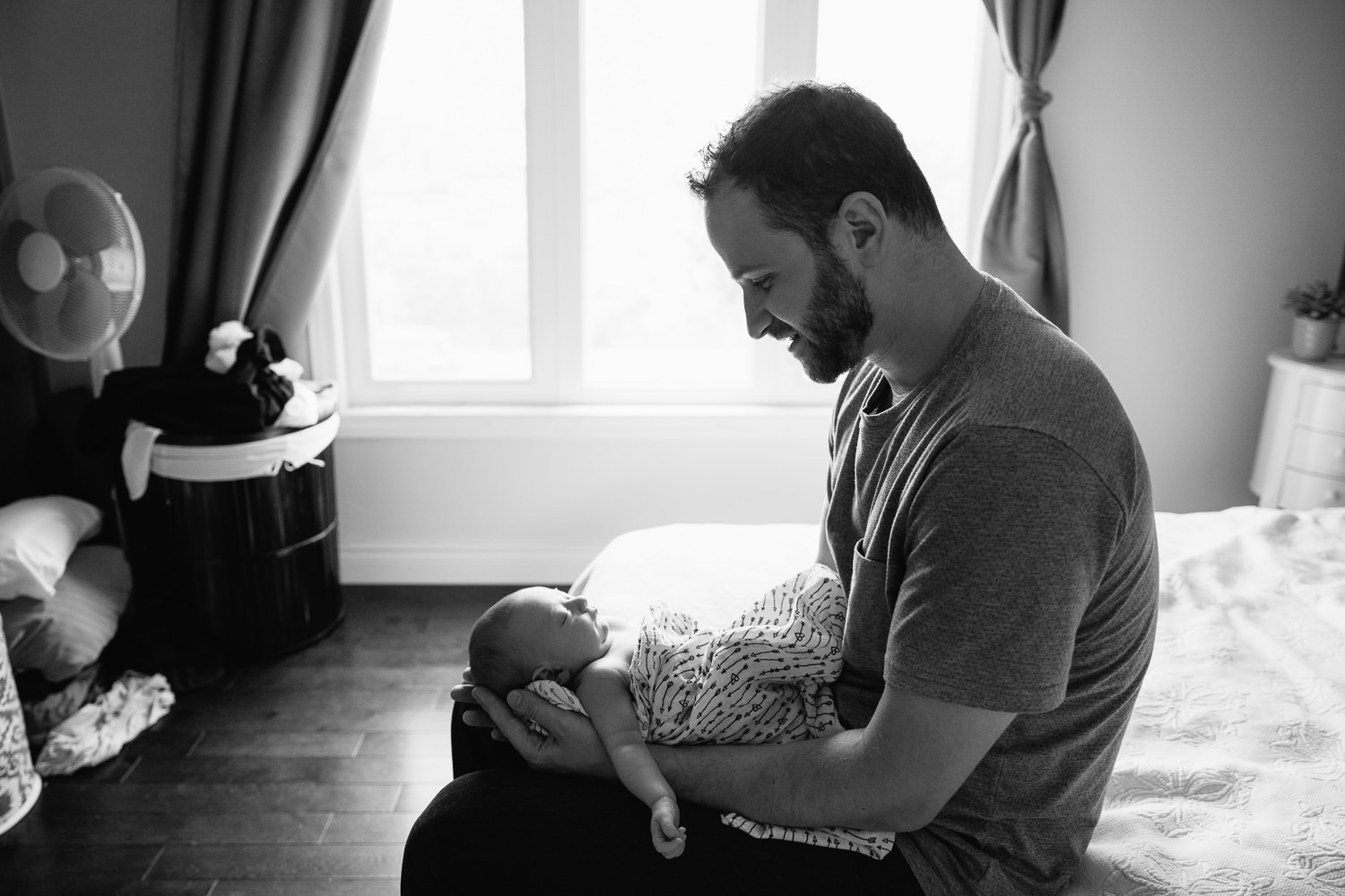 new dad in grey t-shirt sitting on edge of master bed holding sleeping 2 week old baby girl with light hair wrapped in swaddle - York Region In-Home Photos