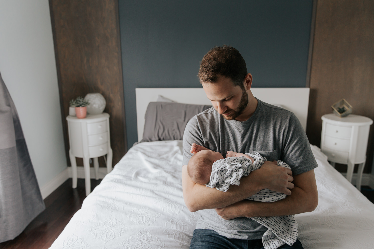 first time father with dark hair and beard in grey shirt sitting on edge of master bed holding sleeping 2 week old baby girl with light hair wrapped in swaddle - Markham In-Home Photos