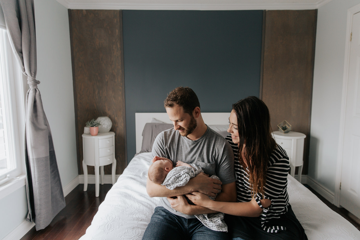 family of 3, new parents sitting on the edge of bed, dad holding 2 week old baby girl with light hair, mom snuggled next to husband, smiling at daughter - York Region In-Home Photography