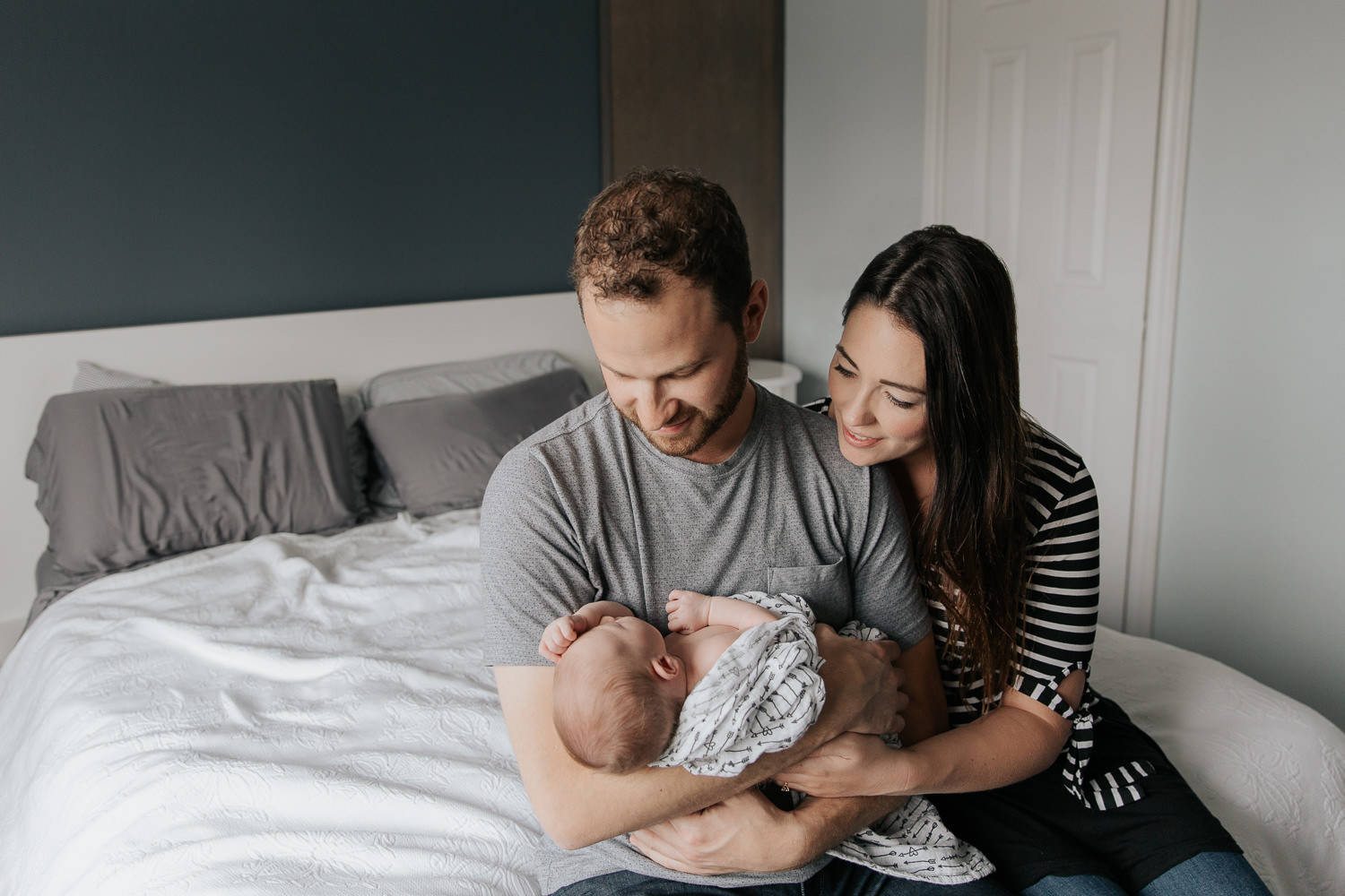 family of 3, new parents sitting on the edge of bed, dad holding 2 week old baby girl with light hair, mom snuggled next to husband, smiling at daughter - Newmarket In-Home Photos