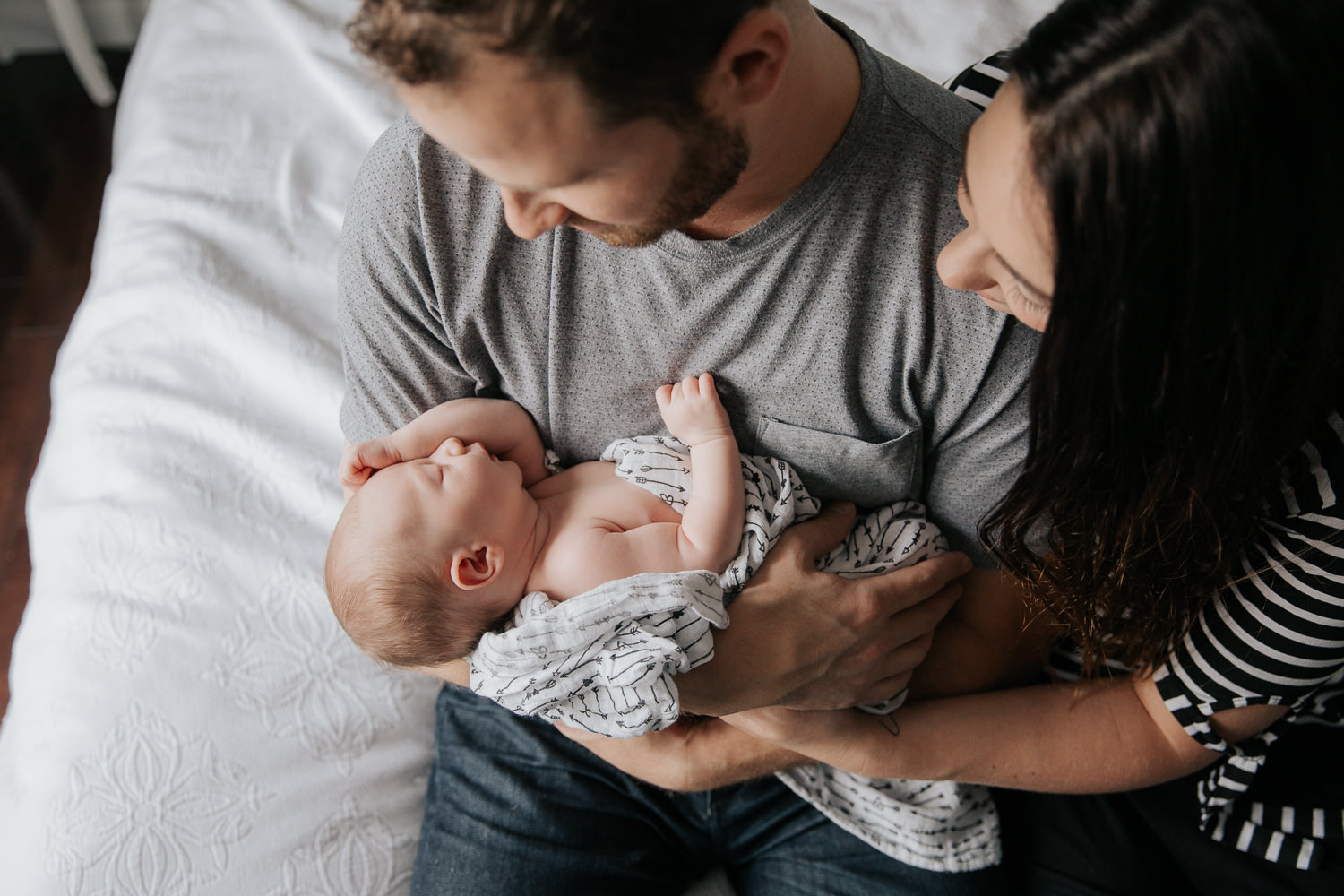 family of 3, new parents sitting on the edge of bed, dad holding 2 week old baby girl with light hair, mom snuggled next to husband, smiling at daughter - Stouffville In-Home Photos