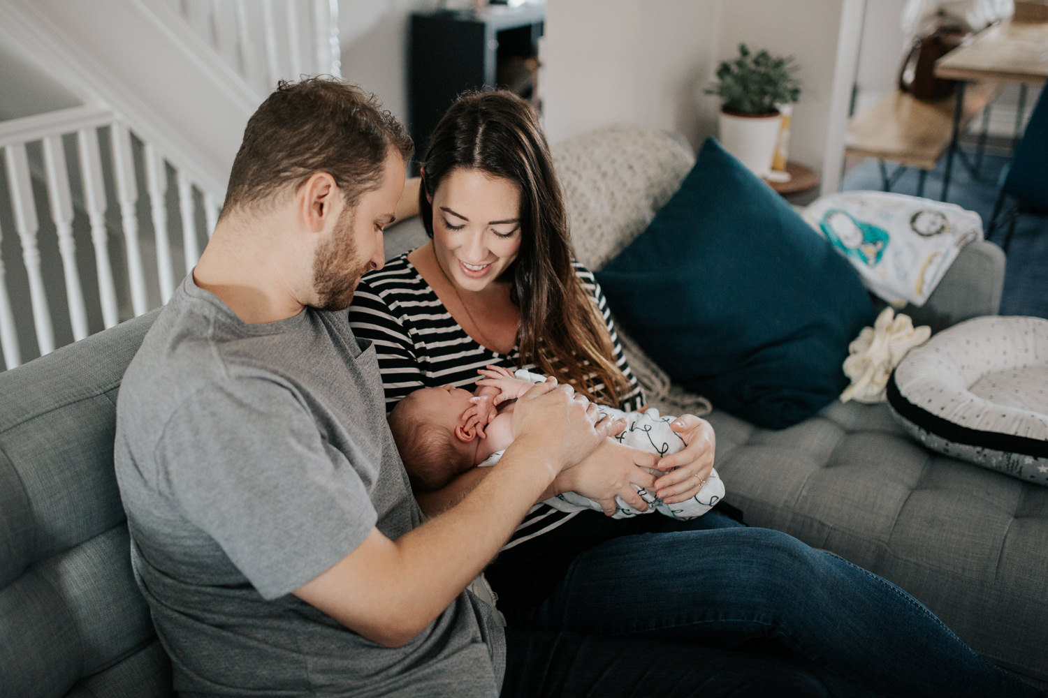 family of 3, new parents cuddle on couch, mom holding 2 week old baby girl covered with black and white swaddle, dad embracing wife and daughter - GTA Lifestyle Photos